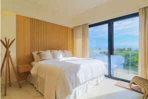 A bedroom with a large bed, a wooden wall panel, a balcony with a view, and a wicker chair. - Luxury 2BR w/ Ocean Views • Gym • Jacuzzi • Infinity Pool • Walk Everywhere (Bucerías)