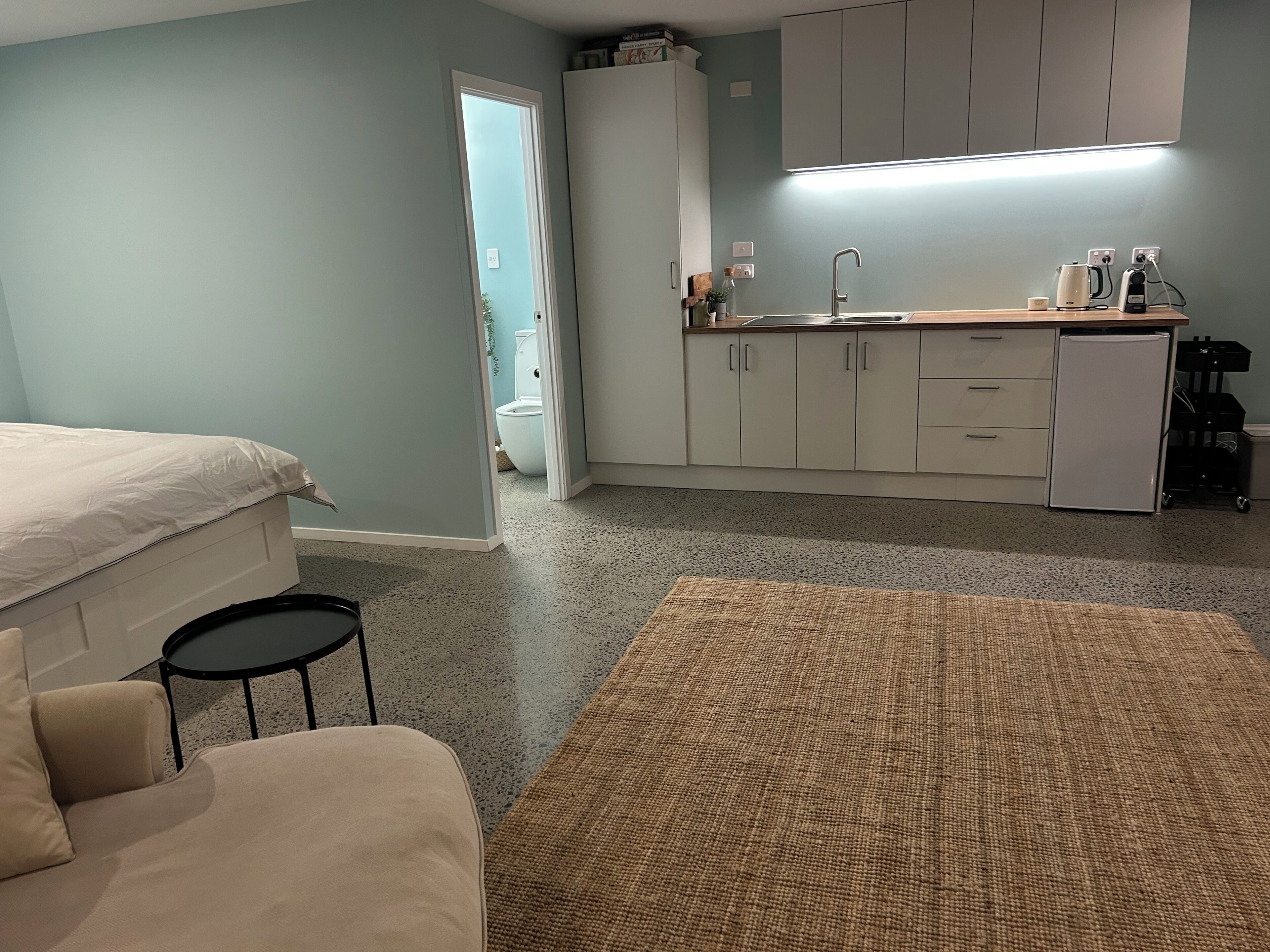 A bedroom with a bed, a small round table, a sofa, a kitchen area with cabinets, a countertop, and a refrigerator.