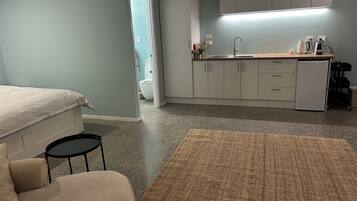 A bedroom with a bed, a small round table, a sofa, a kitchen area with cabinets, a countertop, and a refrigerator.