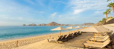 A beach with sun loungers and umbrellas, a clear sky, and distant mountains.