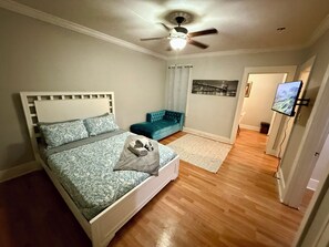 Een slaapkamer met een bed, een blauwe bank, een plafondventilator, een wandtelevisie en een stadsgezicht kunstwerk.