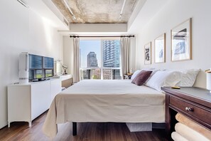 Ein modernes Hotelzimmer mit einem großen Bett, einem Schreibtisch mit Computer, einem Fernseher und einem Fenster mit Blick auf die Stadt.