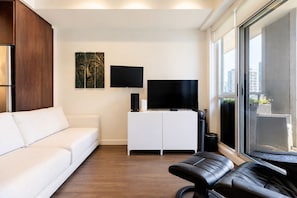 Ein modernes Wohnzimmer mit einem weißen Sofa, einem schwarzen Lederhocker, einer weißen Kommode, einem Flachbildfernseher und einem großen Fenster mit Blick auf die Stadt.