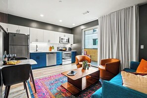Een moderne keuken met witte kastjes, een roestvrijstalen koelkast en een magnetron. Er is een eethoek met een tafel en stoelen, en een zithoek met een bank en salontafel.