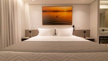 A hotel room with a large bed, two bedside tables with lamps, a painting of a sunset, and a bathroom visible through an open door.