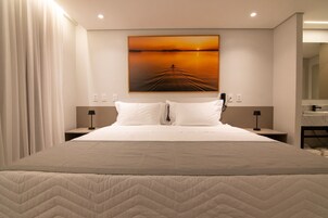 Habitación de hotel con una cama grande, dos mesitas de noche con lámparas, un cuadro de un atardecer y un baño visible a través de una puerta abierta.