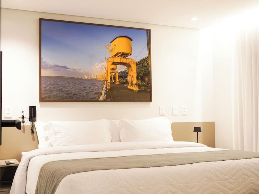 A hotel room with a large bed, a bench, a nightstand, a lamp, and a wall-mounted picture of a coastal scene.