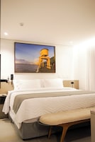 A hotel room with a large bed, a bench, a nightstand, a lamp, and a wall-mounted picture of a coastal scene.