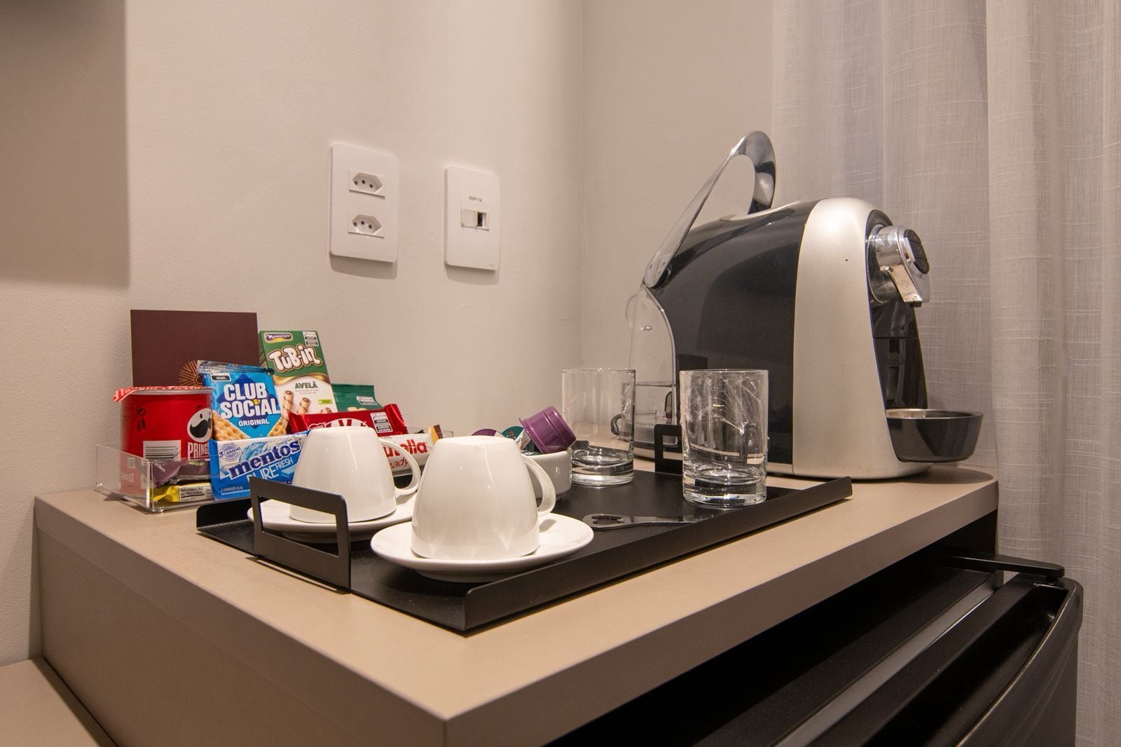 Quarto de hotel com cozinha compacta, cafeteira, chaleira, jarra de café, um copo de água, uma chávena, um prato e uma caixa de cereais Club Social Original.