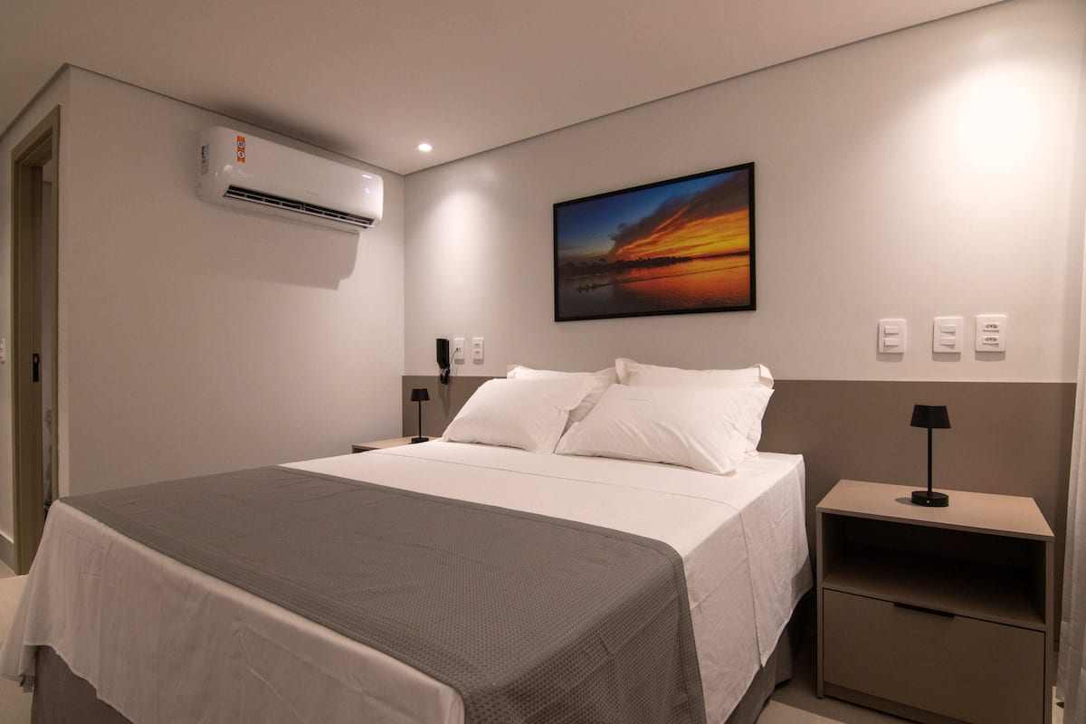 Habitació d'hotel amb llit, taules de nit, un aparell d'aire condicionat, un quadre penjat a la paret i una petita taula al costat del llit.