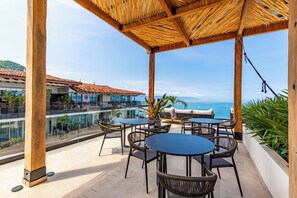 A covered outdoor seating area with tables and chairs, overlooking a pool and a building with a tiled roof. - Suite Estudio de Lujo (Puerto Vallarta)