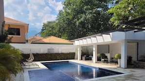 Una piscina con un pergolato e un'area relax, circondata dal verde e da un edificio con tetto in tegole.