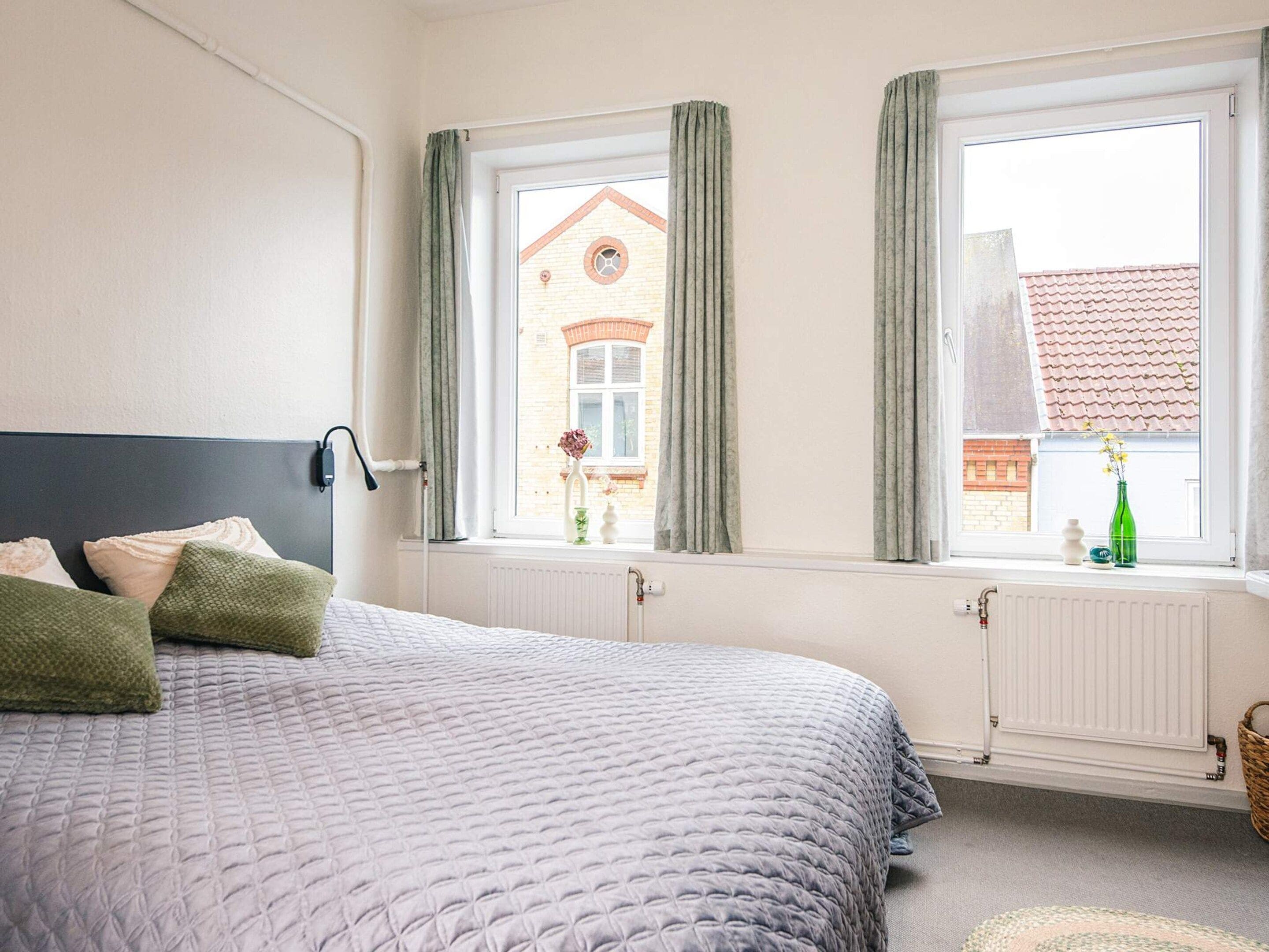 Ein Schlafzimmer mit Bett, einem Fenster mit Vorhängen, einem Heizkörper und Blick auf ein Gebäude.
