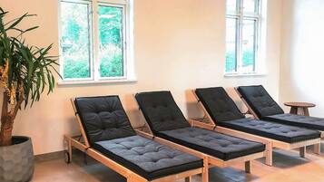 Habitación con cuatro camas lounge de madera con cojines negros, una planta en maceta y ventanas con vistas a vegetación.