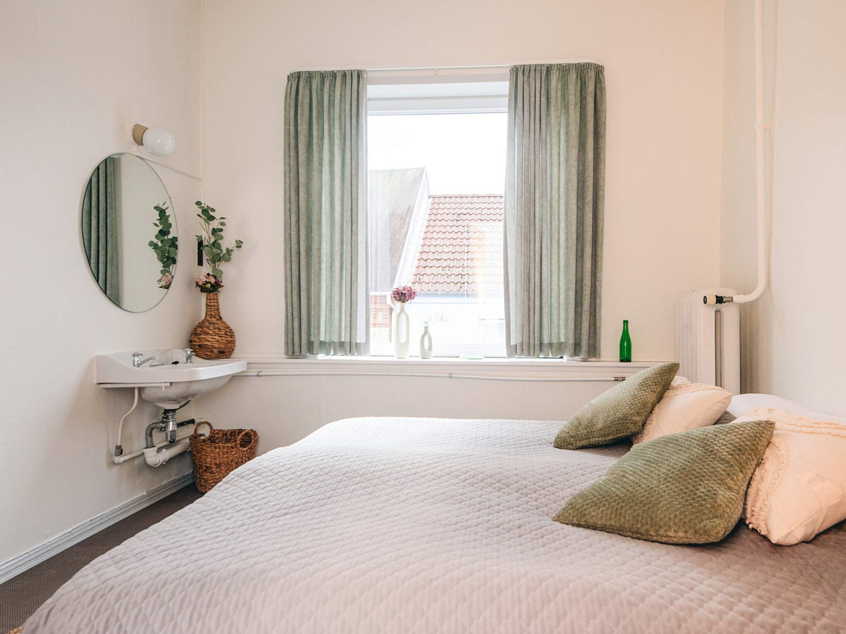A bedroom with a bed, a bedside table, a mirror, a window with curtains, and a radiator.