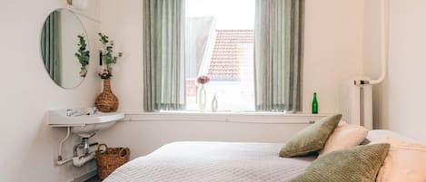 Un dormitorio con cama, mesita de noche, espejo, ventana con cortinas y radiador.