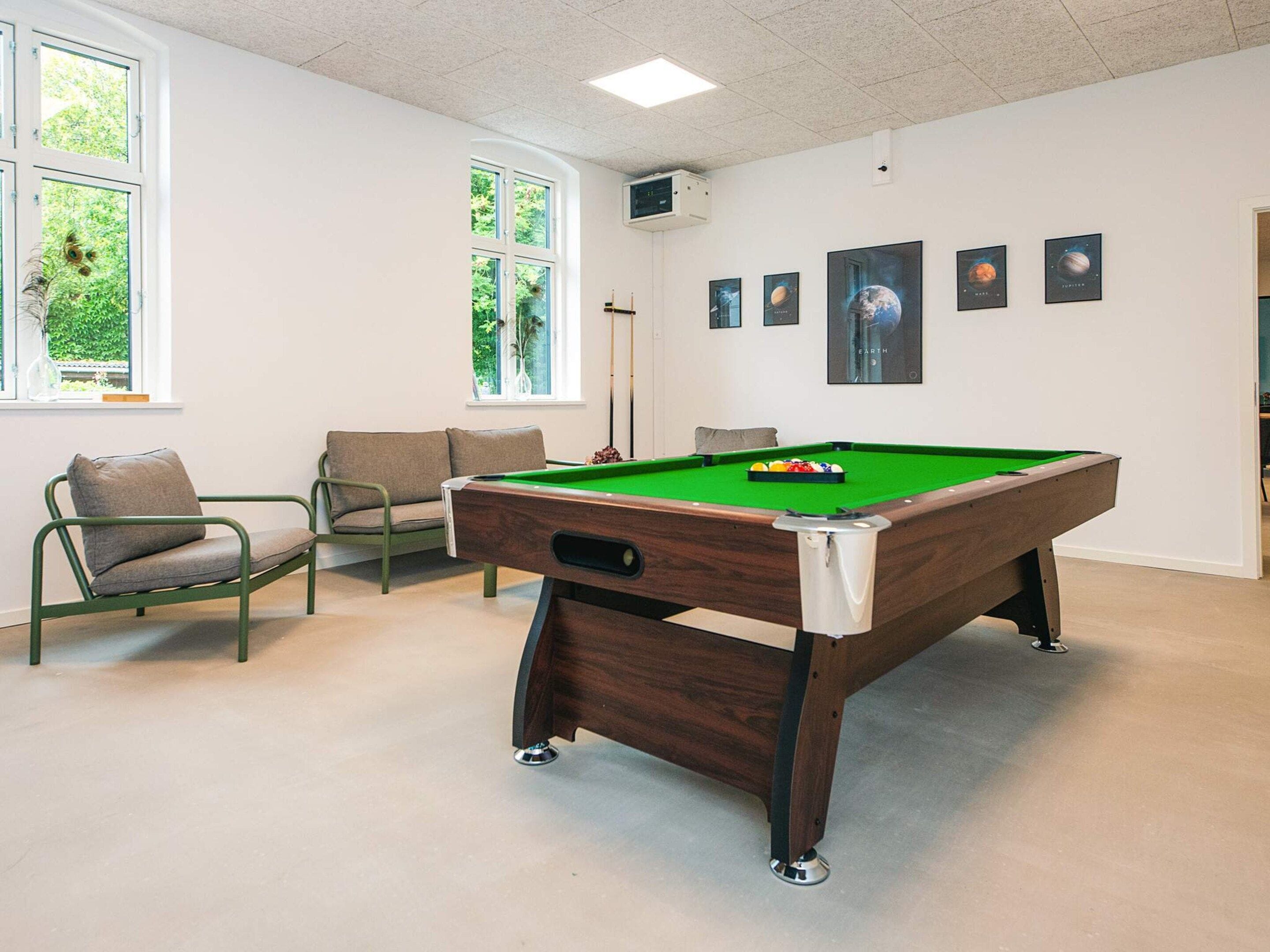 Une table de billard recouverte d’un drap vert, un queue de billard et un jeu de billes. On y trouve deux fauteuils rembourrés, une fenêtre donnant sur un paysage verdoyant et un mur orné de tableaux encadrés.