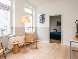 Ein Zimmer mit Holzboden, einem Sessel, einem kleinen Tisch, einer Pendelleuchte und einem Bett im Hintergrund.