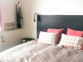 Ein Schlafzimmer mit Bett, Kissen, einem Nachttisch, einem kleinen Beistelltisch, einer Topfpflanze und einer Wandlampe.