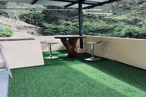 Terraza en la azotea con césped artificial, una mesa de madera en el centro y dos taburetes de bar.