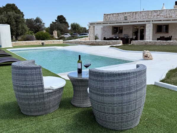 A poolside area with wicker furniture, a bottle of wine, and a stone building in the background. - WHITE STONES HOUSE (Solo Locazione) Trulli e Lamie con piscina e mini spa. (Ostuni)
