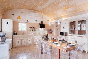 Una cocina con techo de azulejos, sillas transparentes, una mesa de comedor preparada para cuatro personas y un armario blanco con puertas de cristal.