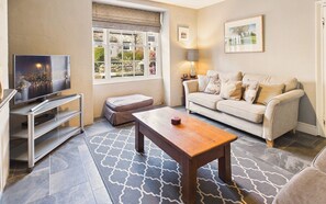 Ein Wohnzimmer mit Fernseher, Sofa, Couchtisch, Hocker und einem Fenster mit Blick auf einen Garten.