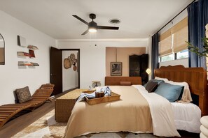 Ein Schlafzimmer mit einem Bett, einem Deckenventilator, einem Nachttisch, einem an der Wand befestigten Regal mit Büchern und einem Fenster mit Vorhängen.