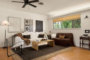 Uma sala de estar com ventilador de teto, janela com vista para árvores, sofá, mesa de centro e abajur.