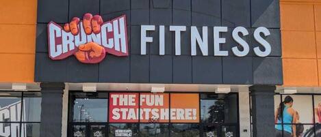 Un gimnasio con un cartel grande que dice 'CRUNCH FITNESS' y el lema '¡LA DIVERSIÓN COMIENZA AQUÍ!'