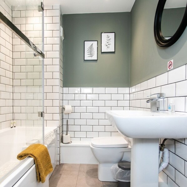 A bathroom with white tiles, a bathtub, a toilet, and a sink. There are framed botanical prints on the wall. - 198 Portland Apartments by Rizalina (Newcastle upon Tyne)