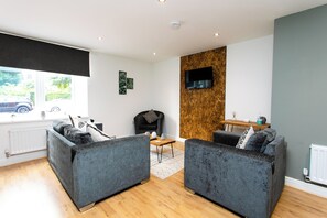 Een moderne woonkamer met grijze banken, een houten accentmuur, een flatscreen televisie en uitzicht op buiten.