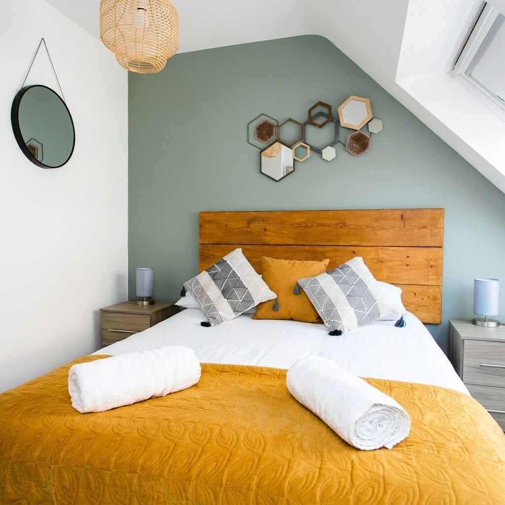 A bedroom with a wooden headboard, a bed with white and yellow bedding, two bedside tables, and a wall-mounted mirror.