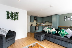 A modern living room with a grey sofa, a wooden coffee table, and a kitchen in the background. - 198 Portland Apartments by Rizalina (Newcastle upon Tyne)