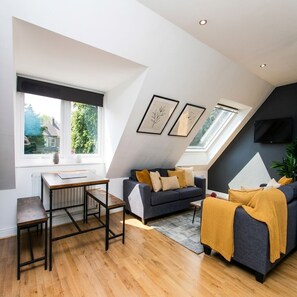 A modern loft apartment with a sloped ceiling, a dining area with a table and chairs, a sofa with cushions, and a television mounted on the wall.
