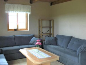 Ein Wohnzimmer mit zwei grauen Sofas, einem Holztisch mit Blumen und einem Fenster mit weißem Vorhang, sowie einem Holzregal.