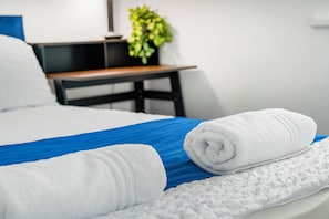 Un lit bien fait, avec une couvre-lit bleue et du linge de lit blanc, une table de chevet en bois ornée d’une plante, et un mur blanc.