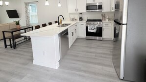Una cucina moderna con mobili bianchi, elettrodomestici in acciaio inossidabile, un'isola centrale, illuminazione a sospensione e un'area pranzo con tavolo e sedie scure.