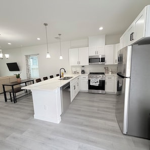 A modern kitchen with white cabinetry, stainless steel appliances, a central island, pendant lighting, and a dining area with a dark table and chairs. - New Construction 3-bedroom house in vibrant Houston (Houston)