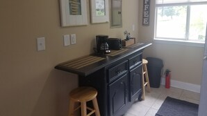 A kitchen with a black countertop, wooden stools, a coffee sign, and framed pictures on the wall. - Cozy studio condo in Biloxi. Walk to the beach! (Biloxi)