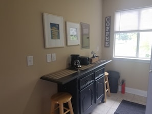 A kitchen with a black countertop, wooden stools, a coffee sign, and framed pictures on the wall. - Cozy studio condo in Biloxi. Walk to the beach! (Biloxi)