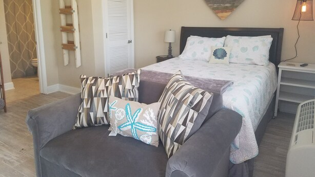 A bedroom with a bed, a nightstand, a chair, a heart-shaped wall decoration, and a ceiling fan. - Cozy studio condo in Biloxi. Walk to the beach! (Biloxi)