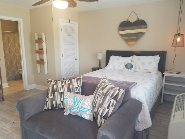 A bedroom with a bed, a nightstand, a chair, a heart-shaped wall decoration, and a ceiling fan. - Cozy studio condo in Biloxi. Walk to the beach! (Biloxi)