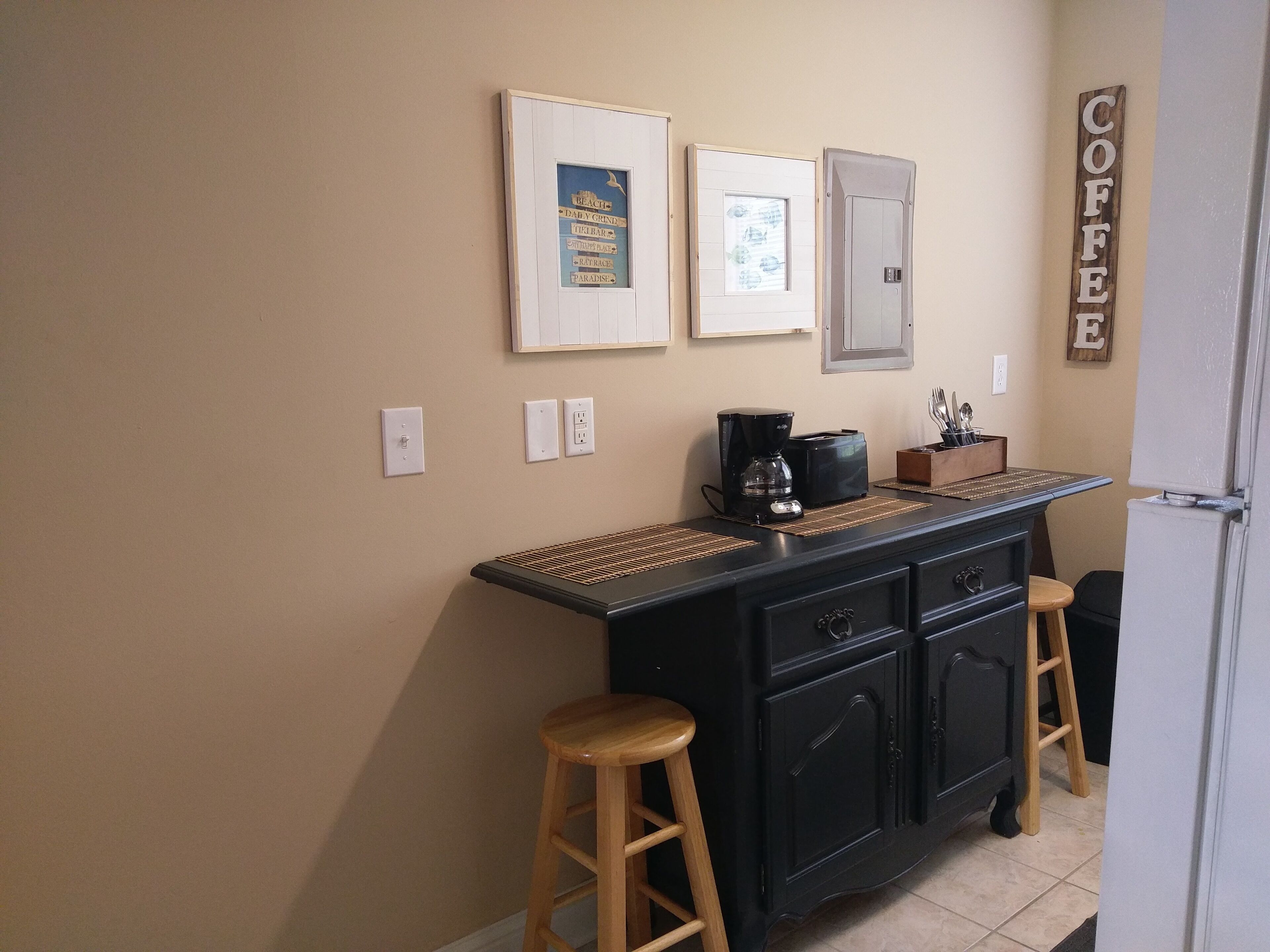 A kitchen with a coffee station, framed signs on the wall, and a refrigerator.