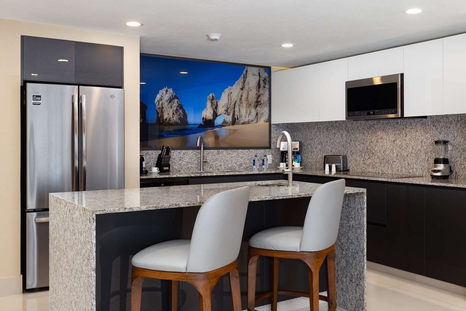 A modern kitchen with a granite countertop, stainless steel appliances, and a large TV mounted above the sink.