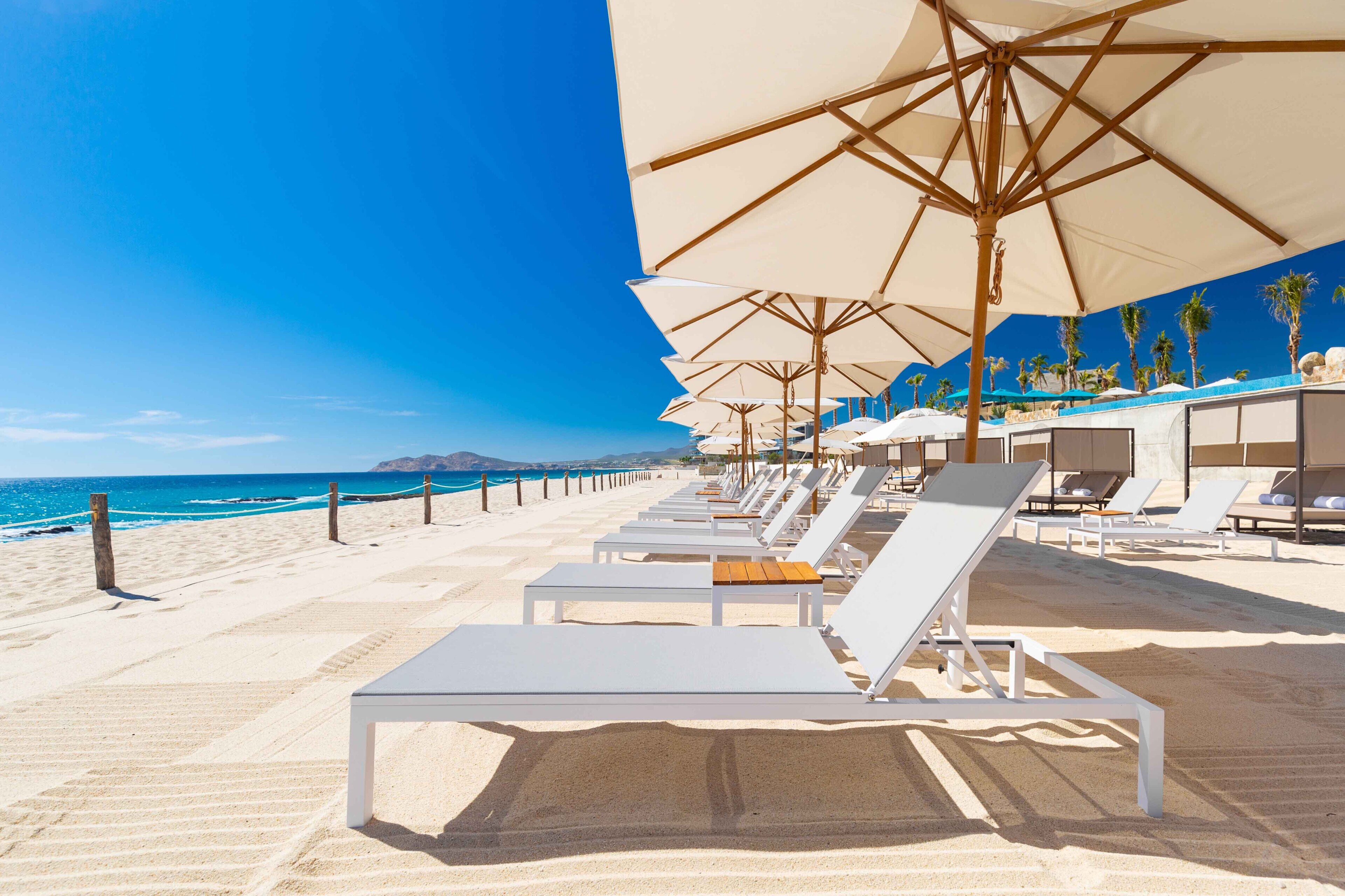 A beachside hotel with sun loungers and umbrellas, a clear blue sky, and the ocean in the background.
