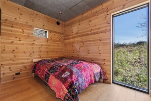 A room with wooden walls, a bed with a colorful quilt, a small window, and a view of greenery outside. - Charming tiny house in forest near sea peace now (Hasle)