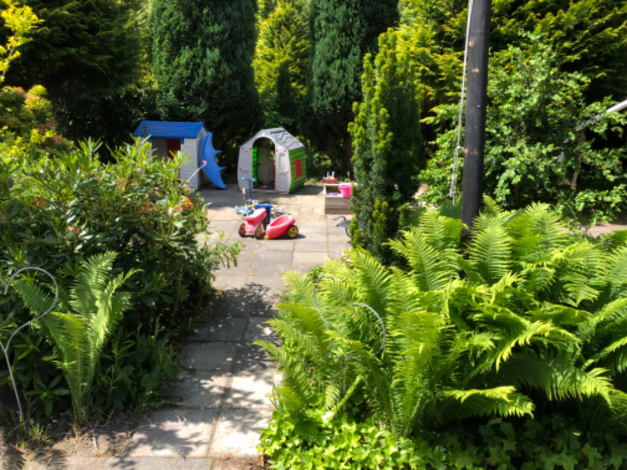 A garden pathway with a blue playhouse, a white archway, and a red and white play vehicle.