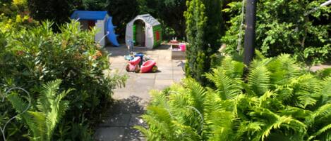 A garden pathway with a blue playhouse, a white archway, and a red and white play vehicle.