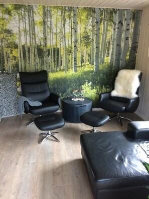 Ein Zimmer mit einem großen Fenster, einem schwarzen Ledersofa, einem schwarzen Leder-Sessel und einem kleinen runden Tisch mit einer weißen, flauschigen Decke.